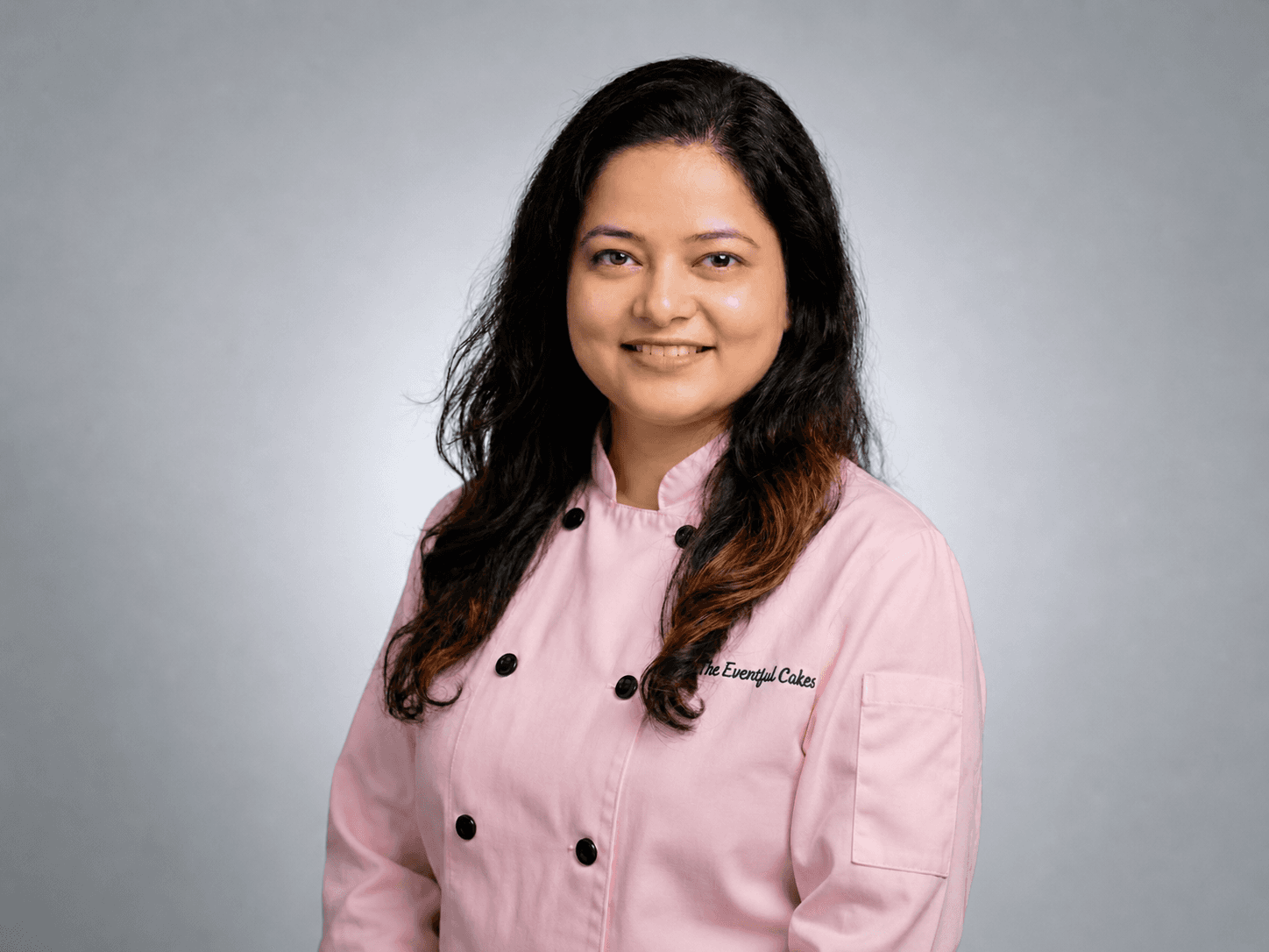 Smiling woman with long dark hair wearing a pink chef's jacket from The Eventful Cakes.